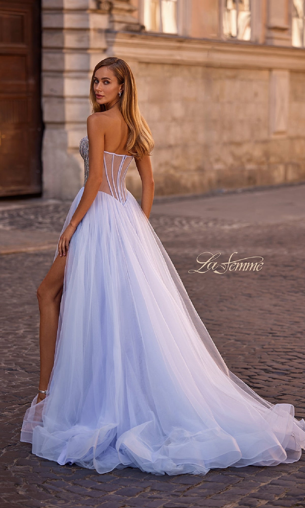 A woman in the La Femme 32811 Strapless Long Prom Ball Gown—white tulle, beaded sweetheart corset bodice, thigh-high slit—stands on a cobblestone street, looking over her shoulder.